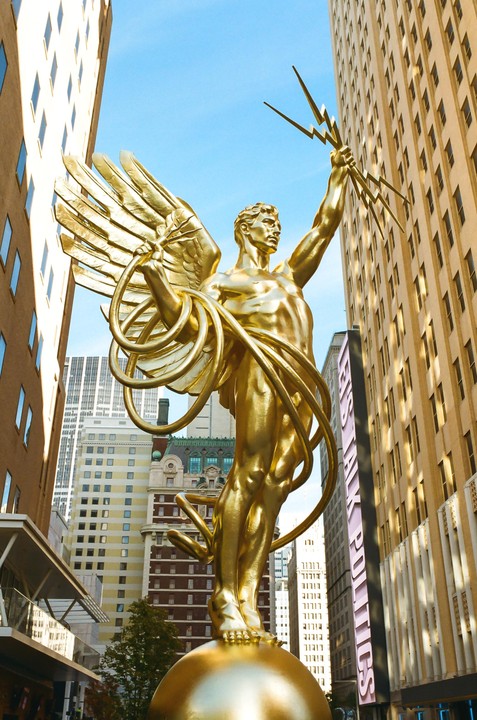 A golden statue of a winged man holding lightning bolts stands tall between two buildings in a city.