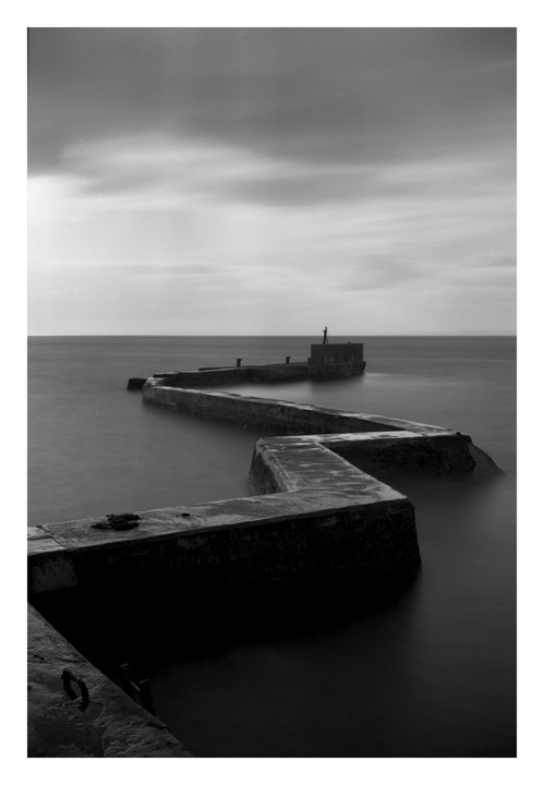 A zig-zag breakwater extends into the ocean under a cloudy sky.