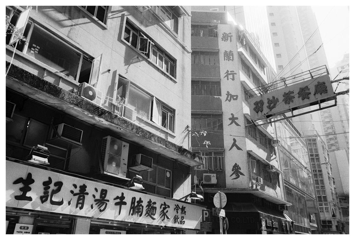 A street in Hong Kong is lined with buildings featuring signs with Chinese characters and numerous air conditioning units.