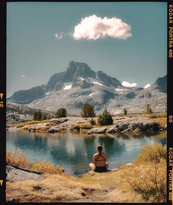 A woman sits by a lake with mountains in the background.