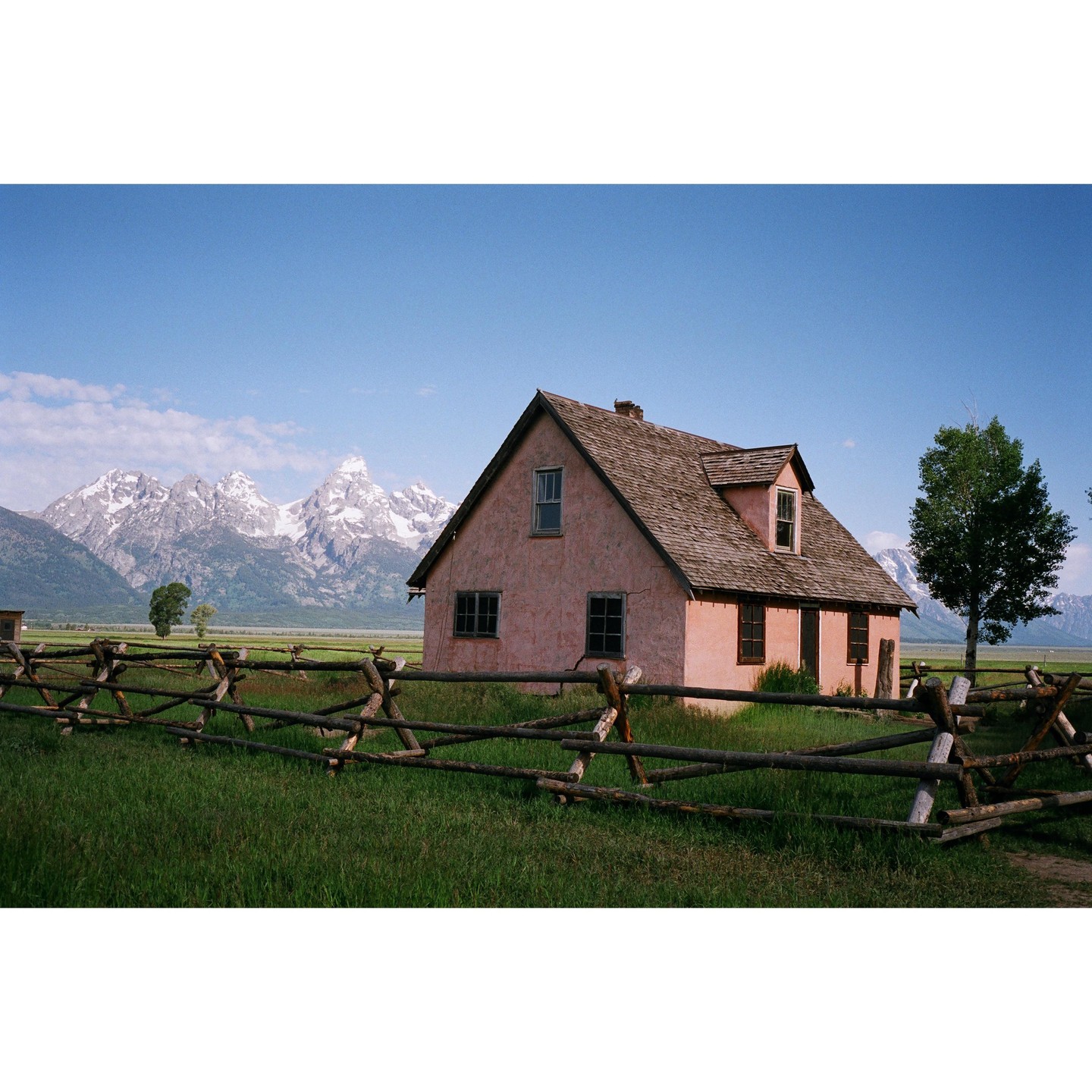A pink house with a wooden fence in the foreground stands before a mountain range with snow-capped peaks under a clear blue sky.