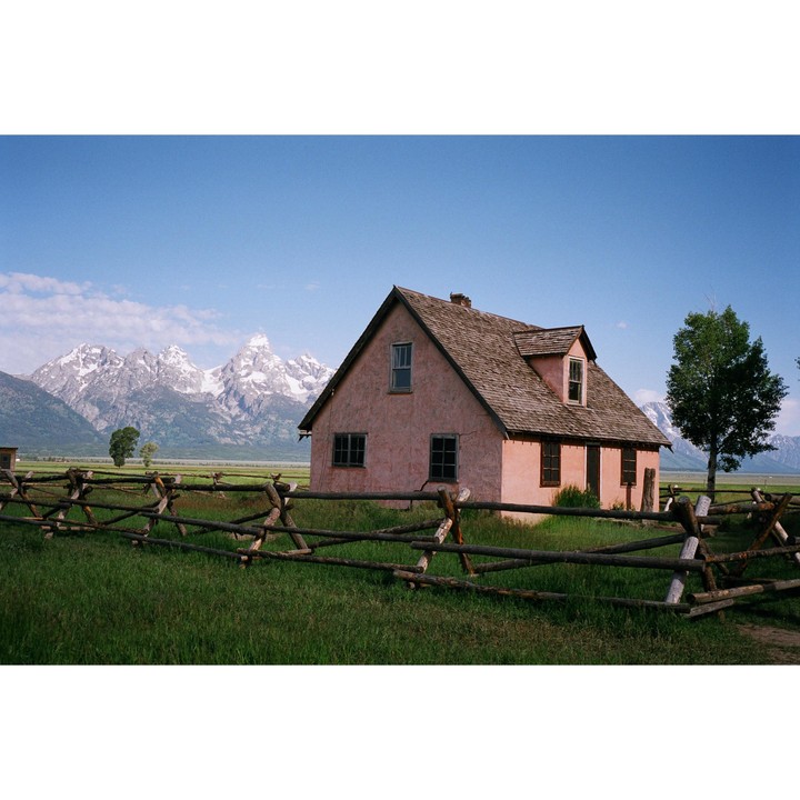 A pink house with a wooden fence in the foreground stands before a mountain range with snow-capped peaks under a clear blue sky.