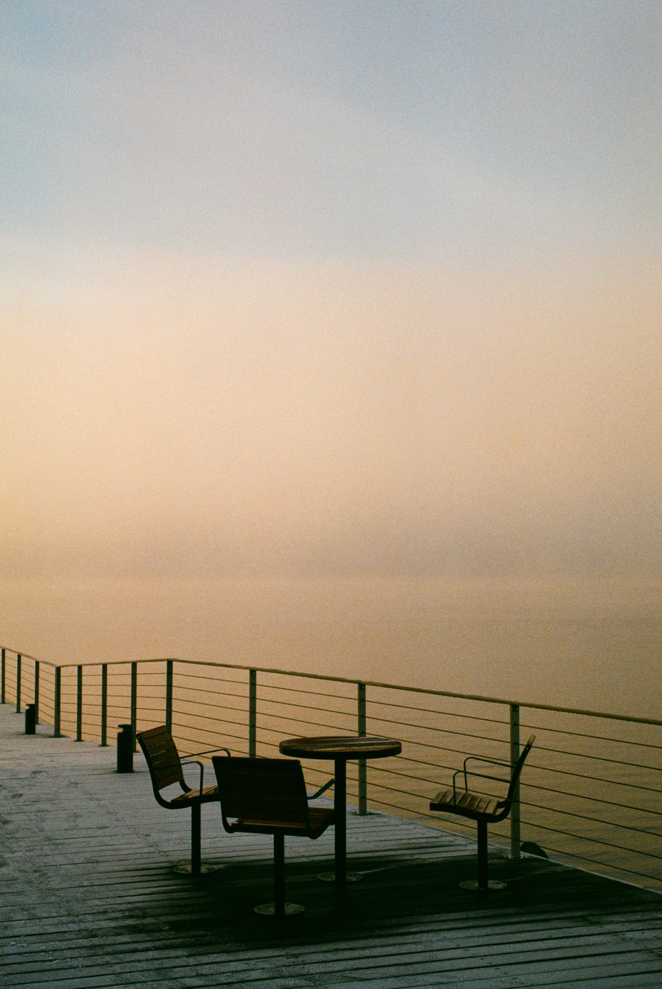 A table and two chairs sit on a pier overlooking a hazy body of water under a pale sky.