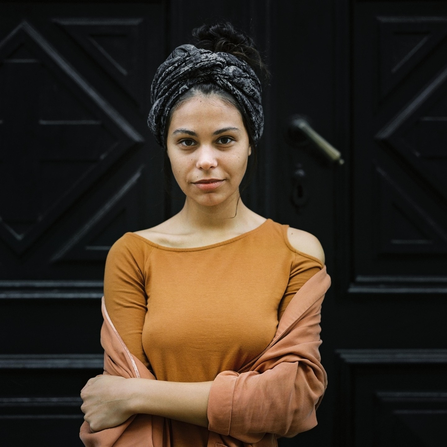 A woman with a head wrap and a brown shirt stands with her arms crossed in front of a dark door.