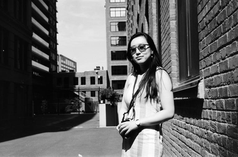 A woman wearing sunglasses stands next to a brick wall with buildings in the background.