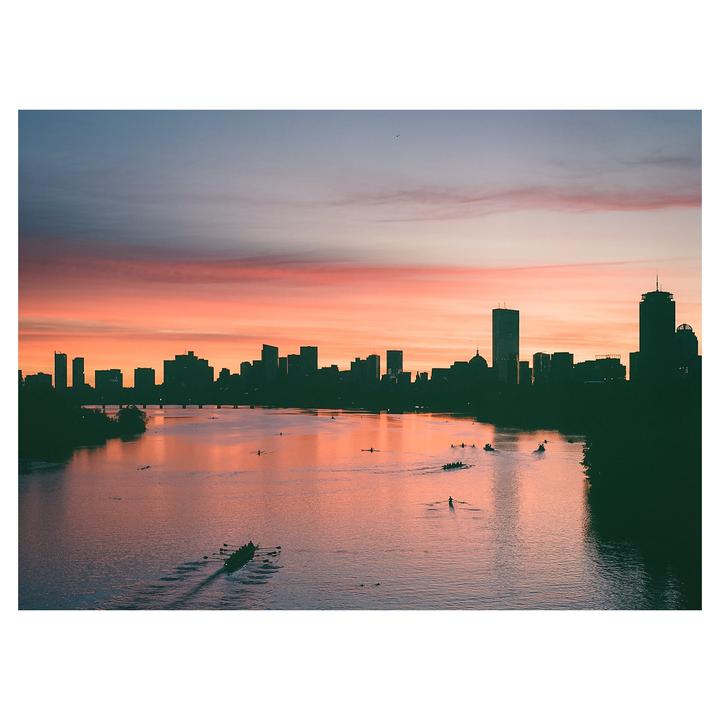 A city skyline is silhouetted against a colorful sunrise sky over a river with several rowing boats.