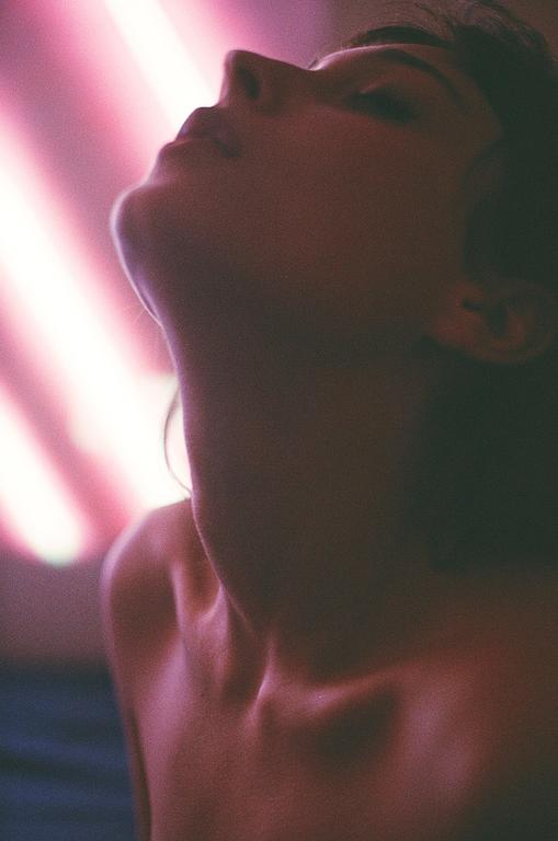 A woman's head is tilted back, with her face and neck illuminated by pink light.