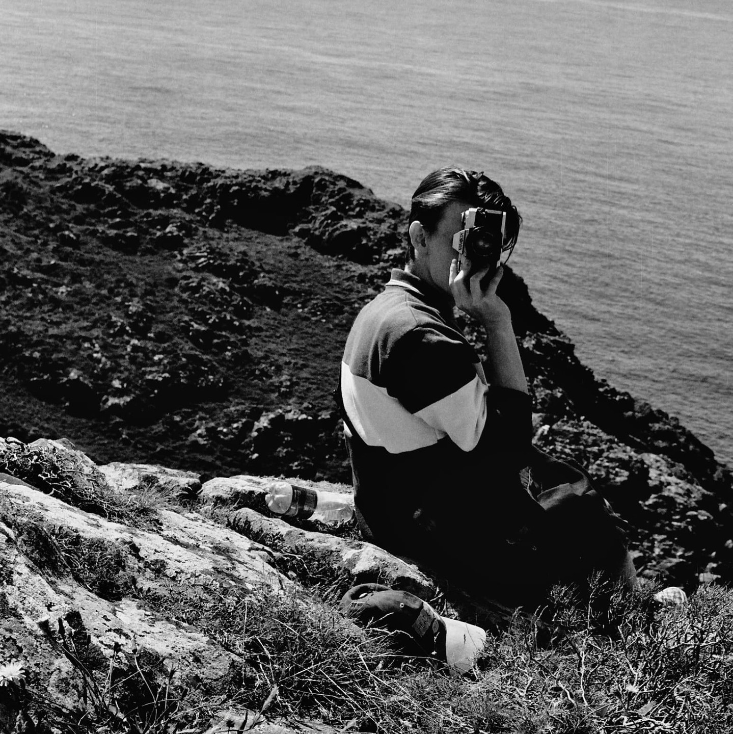 A man sits on a cliff overlooking the ocean and takes a photo.