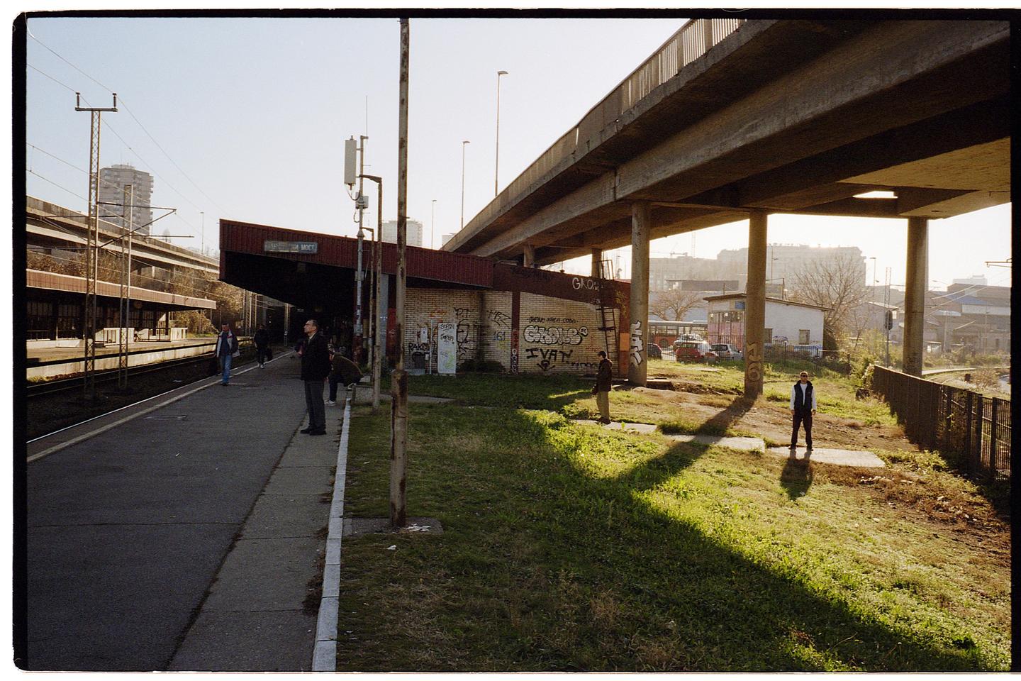 A man stands on a grassy slope under a concrete overpass next to a train station platform where other people are waiting.