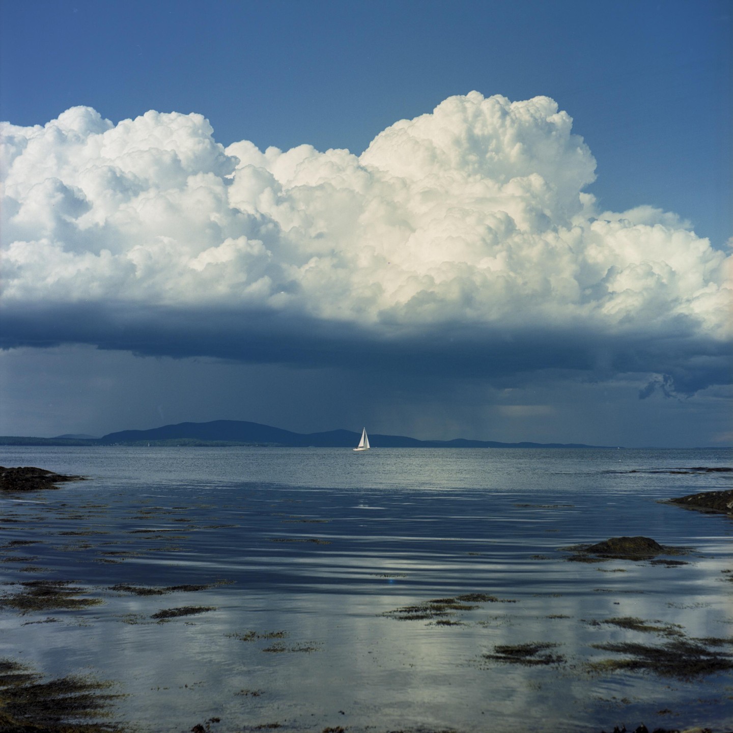 A sailboat is visible on the water under a large cloud formation with a dark, stormy base.