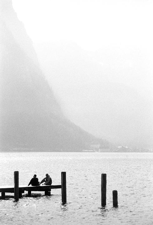 Two people sit on a dock extending into a lake with a hazy mountain in the background.