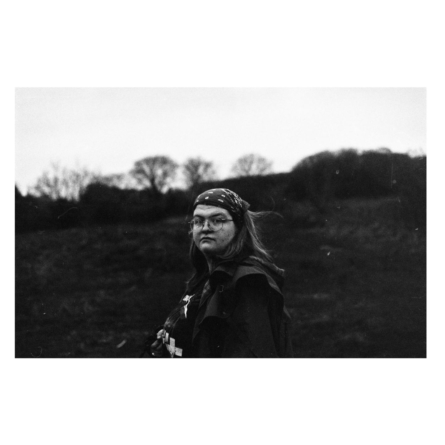 A woman wearing glasses and a bandana looks at the camera in a field with trees in the background.