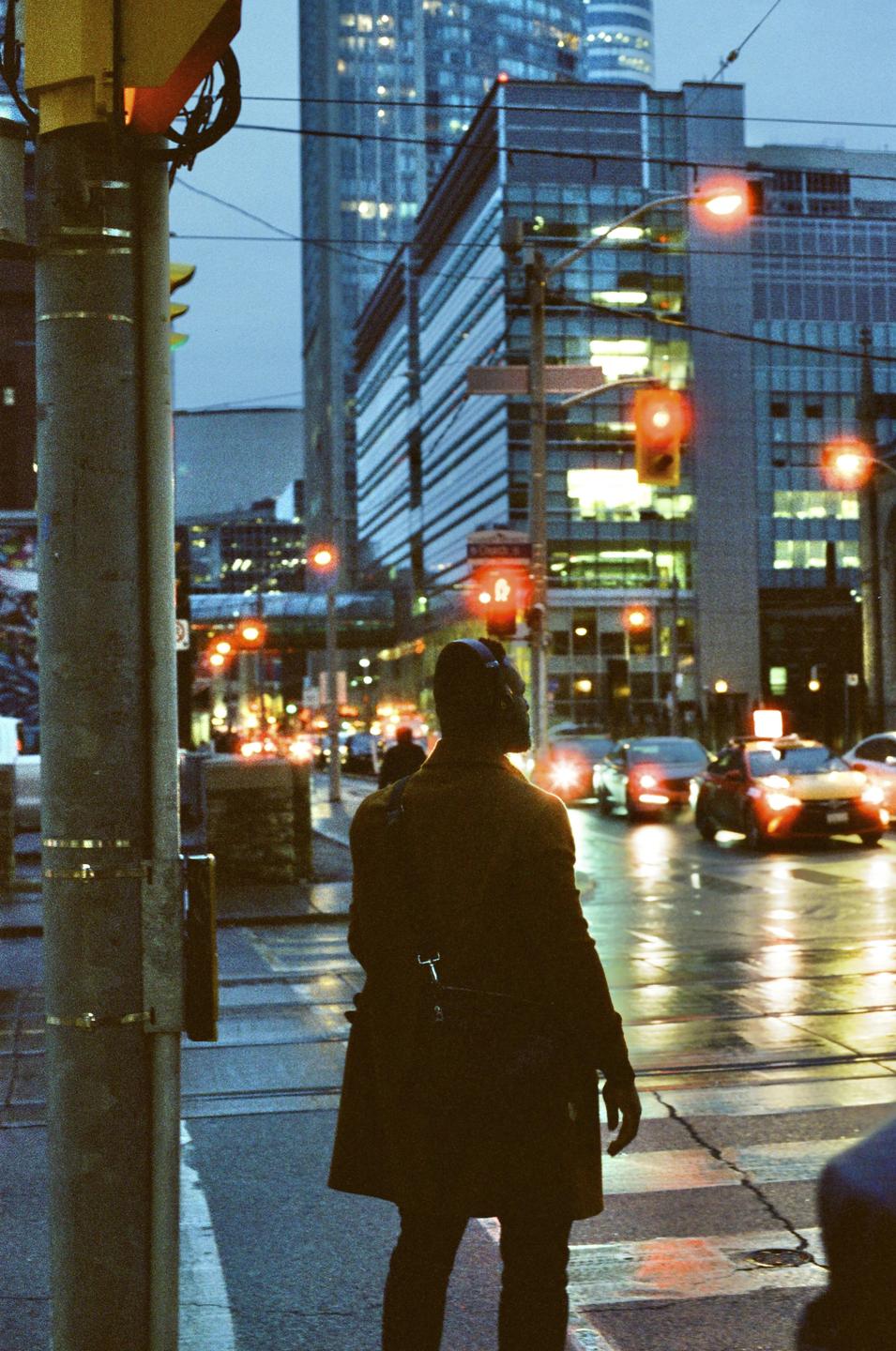 A man stands at a crosswalk in a city at dusk, with cars and buildings visible in the background.