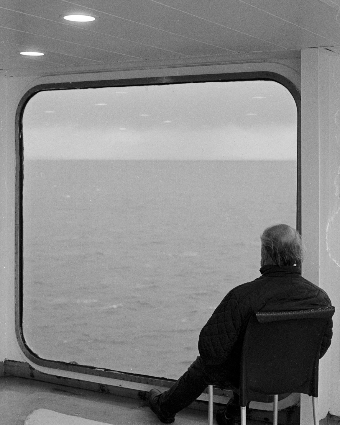 A man sits on a chair looking out a large window at the sea.
