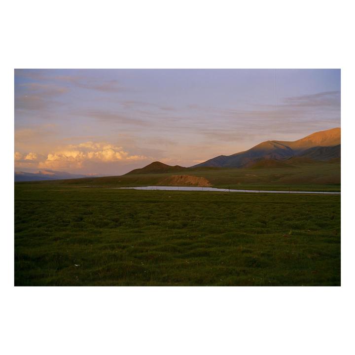A vast green field stretches towards distant mountains under a sky with sunset clouds.