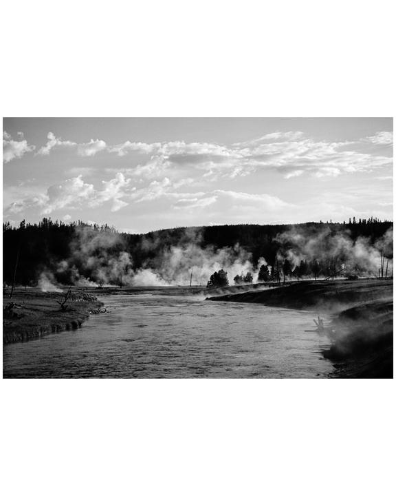 A river flows through a landscape with steam rising from the banks and a forest in the background under a cloudy sky.