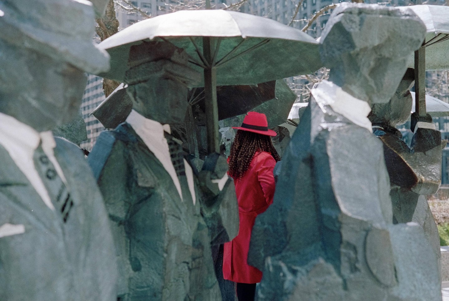 A woman in a red coat and hat stands among several statues of men holding umbrellas.