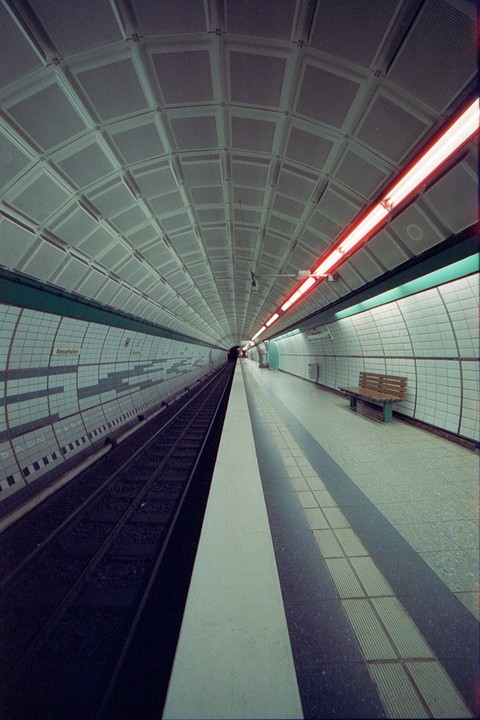 A long, empty subway platform stretches into the distance with tracks on the left and a bench on the right.
