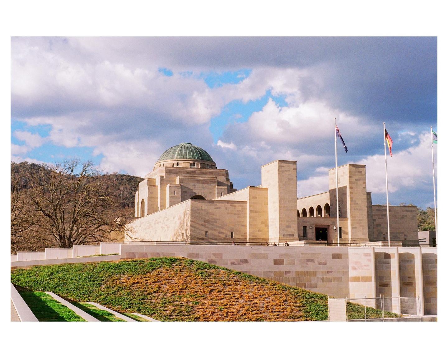 A large stone building with a green dome stands under a partly cloudy sky, with flags flying in front and a hillside in the background.