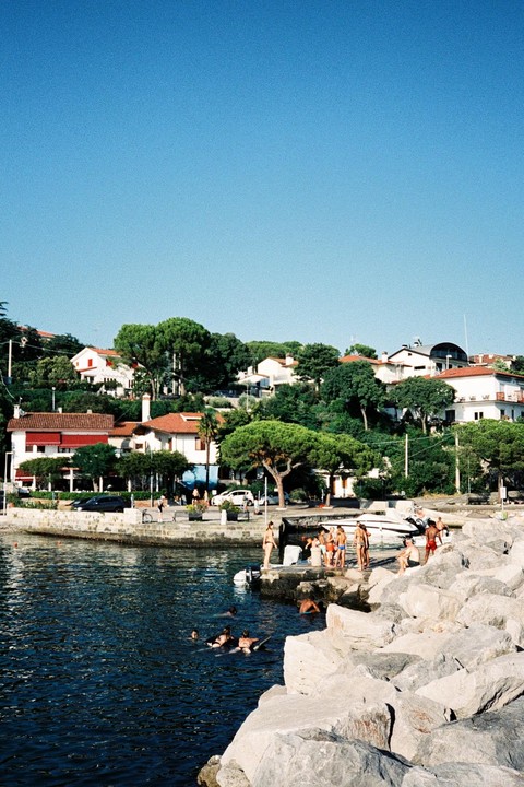 People swim in the water and relax on a rocky shore with houses and trees on a hill in the background.