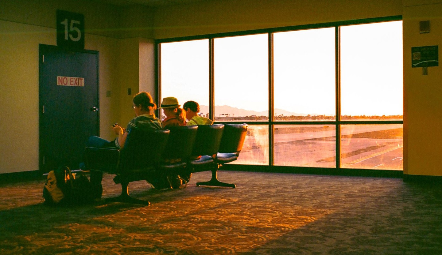 Three people sit in an airport waiting area looking out a large window at an airfield during golden hour.