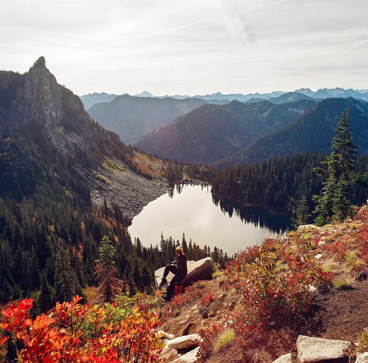 A woman sits on a rock overlooking a lake surrounded by mountains and autumn foliage.