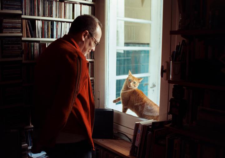 A man in a red jacket looks at an orange cat sitting on a window sill.