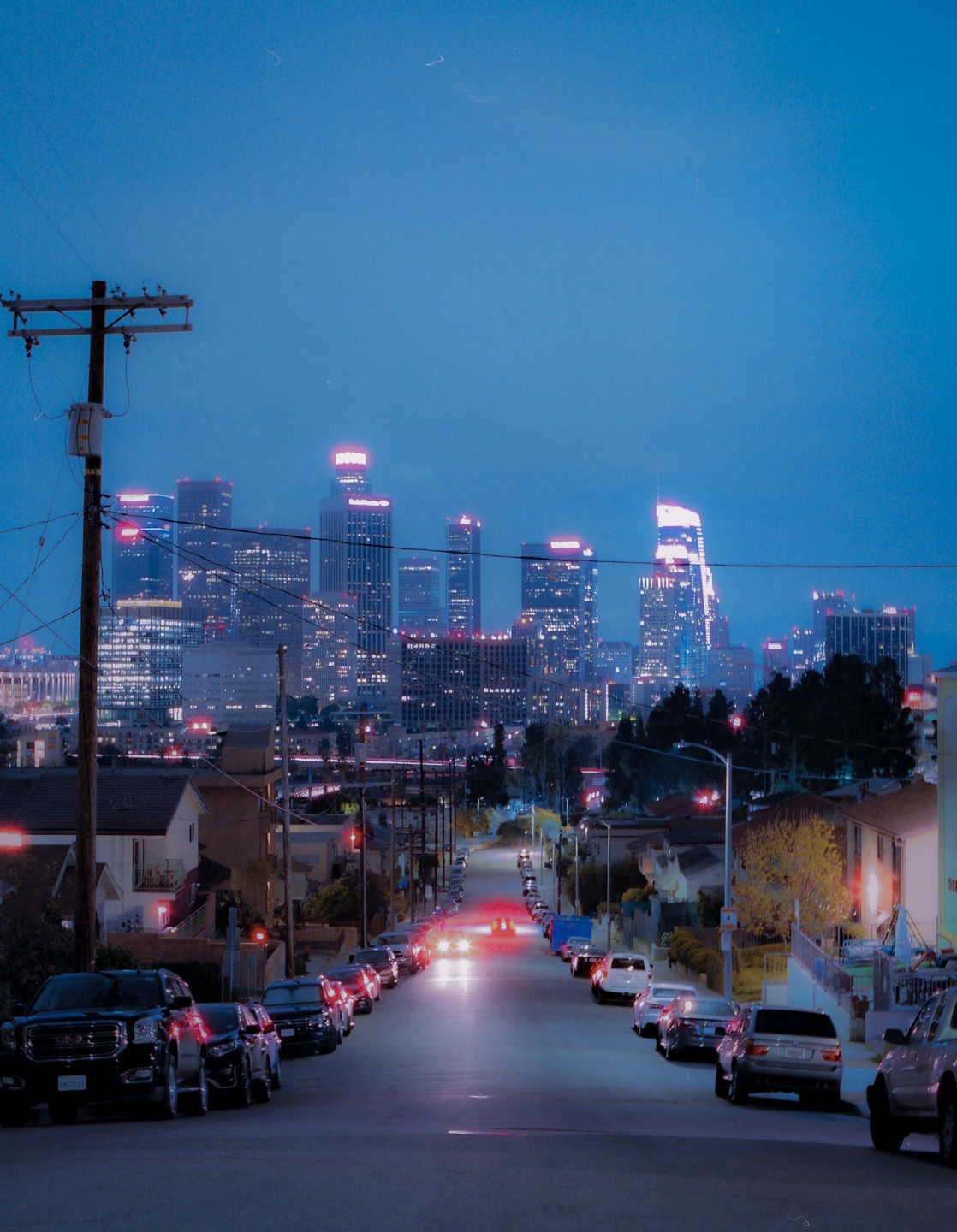 A street lined with parked cars leads towards a distant city skyline at night.