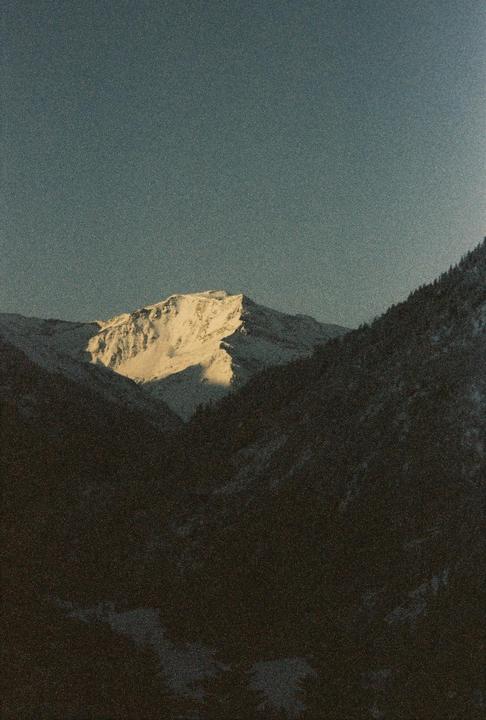 A snow-covered mountain peak is illuminated by sunlight, contrasting with the dark, shadowed mountains in the foreground.