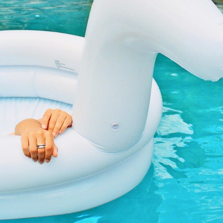 A person's hands rest on the edge of a white inflatable pool float in a swimming pool.