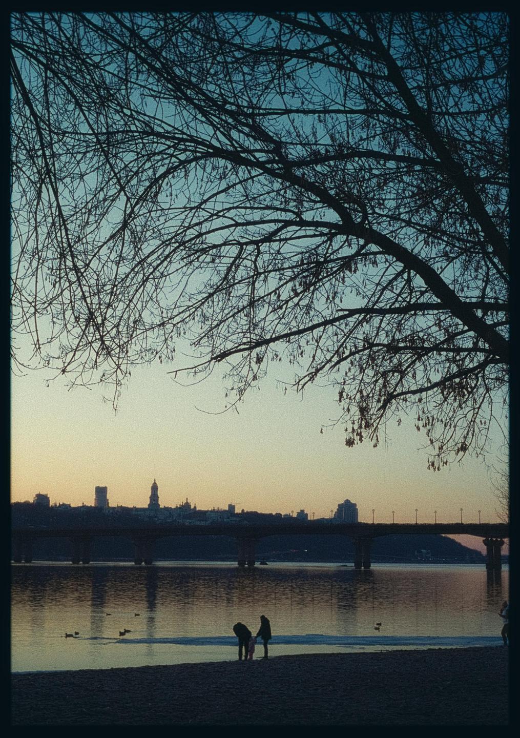 Two people stand on a beach next to a river with a city skyline and bridge in the background, framed by tree branches.