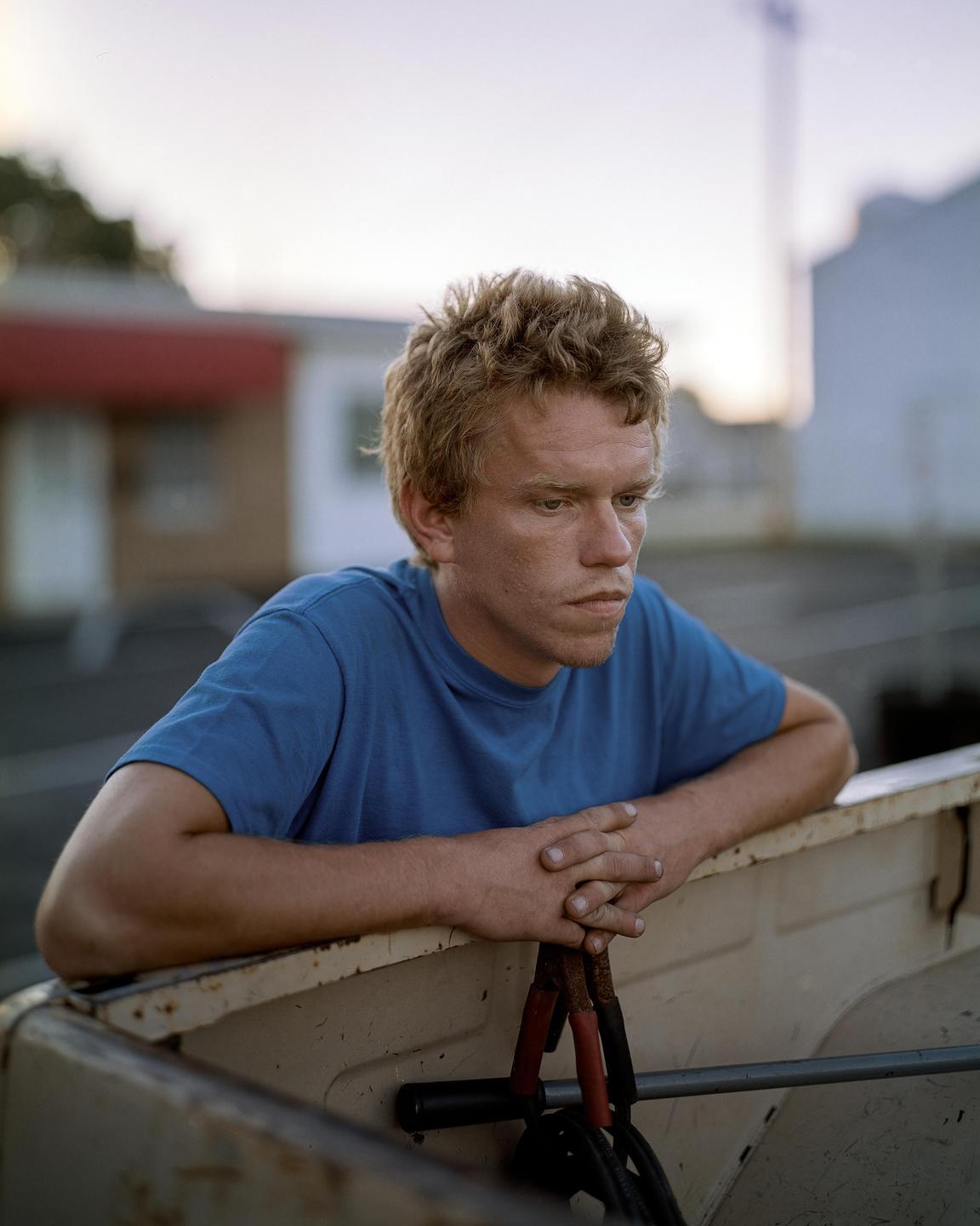 A man with red hair and a blue shirt leans on the back of a truck with jumper cables.