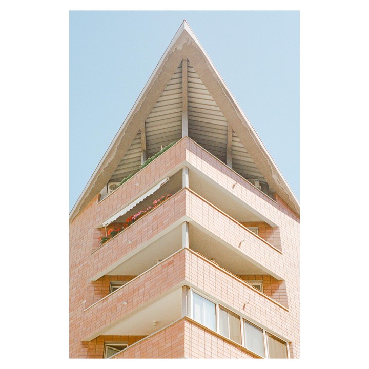 A building with a triangular roof and balconies is shown from a low angle against a clear sky.