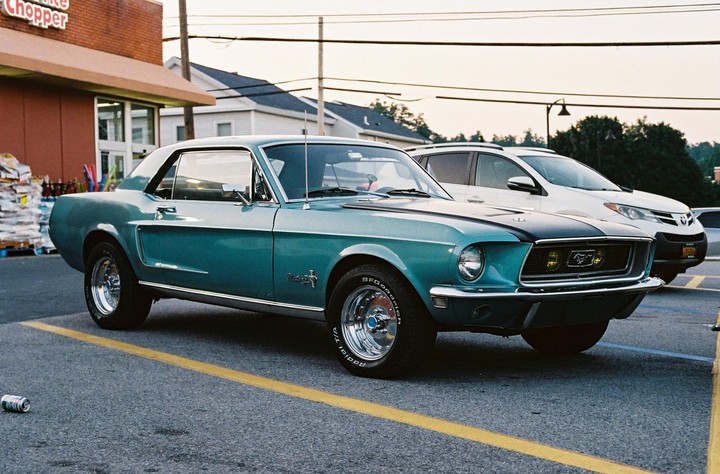 A teal vintage Mustang is parked in a parking lot next to a white car and a building.