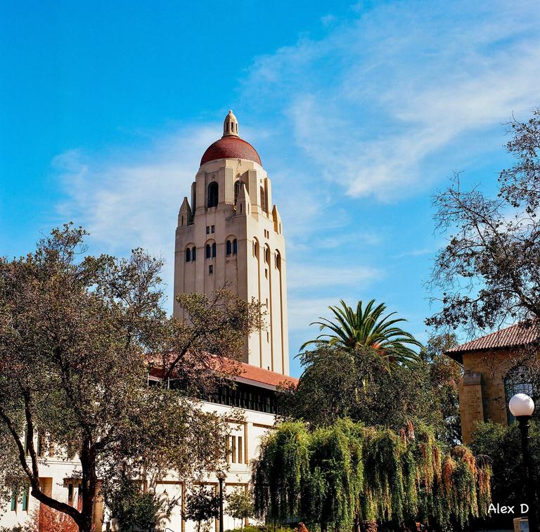 A tall, light-colored tower with a red dome rises above trees and buildings under a blue sky.