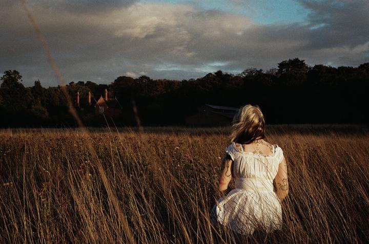 A woman in a white dress stands in a field of tall grass, looking towards a house in the distance under a cloudy sky.