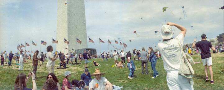 A large crowd of people are gathered on a grassy field with the Washington Monument and many American flags in the background, and kites flying in the sky.