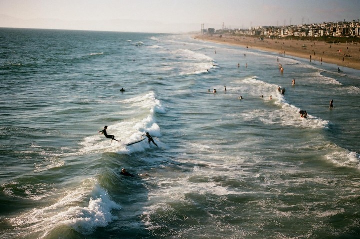 A wide shot shows surfers and swimmers in the ocean next to a beach with buildings in the distance.