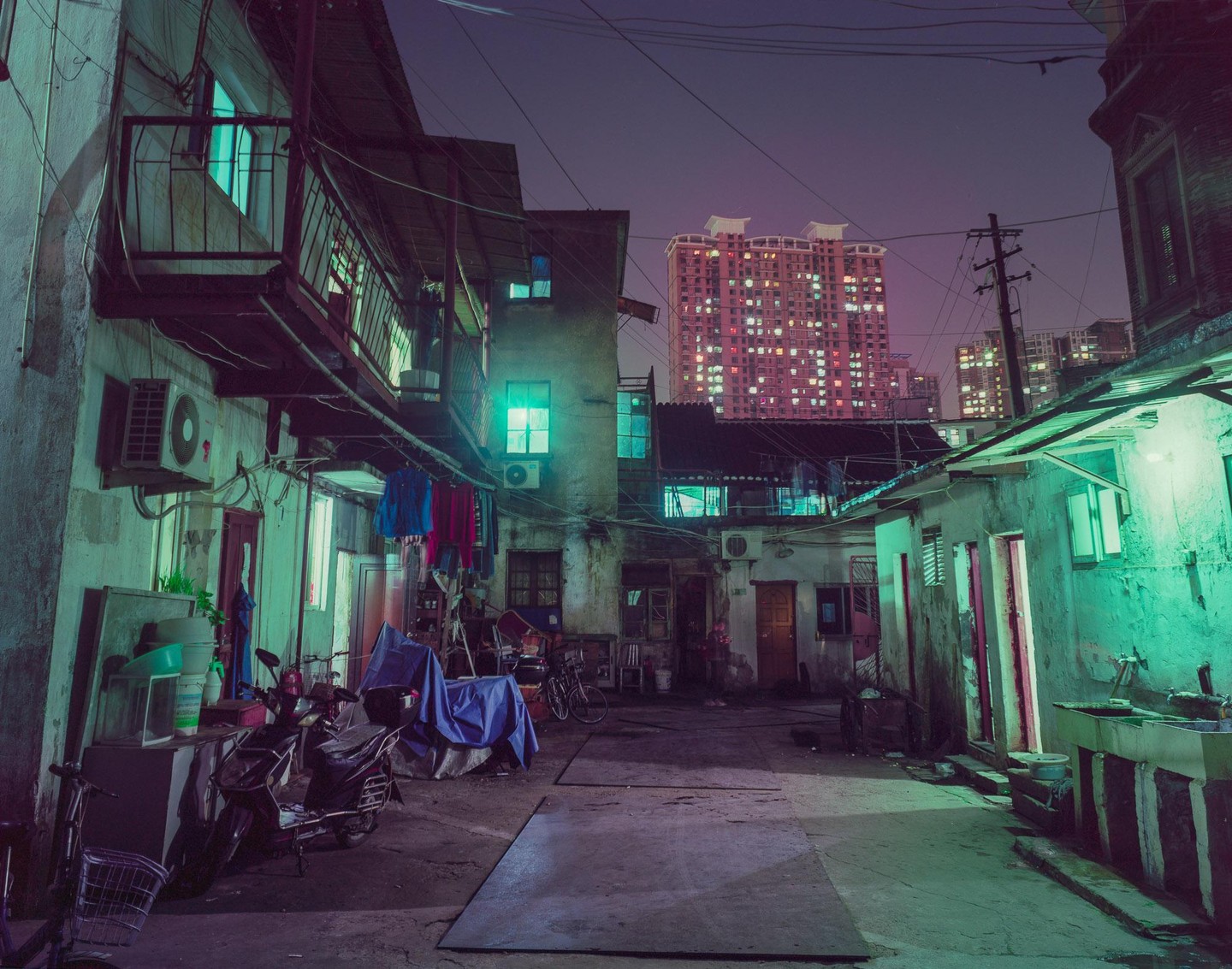 A narrow alleyway at night is illuminated by green and pink light from surrounding buildings.