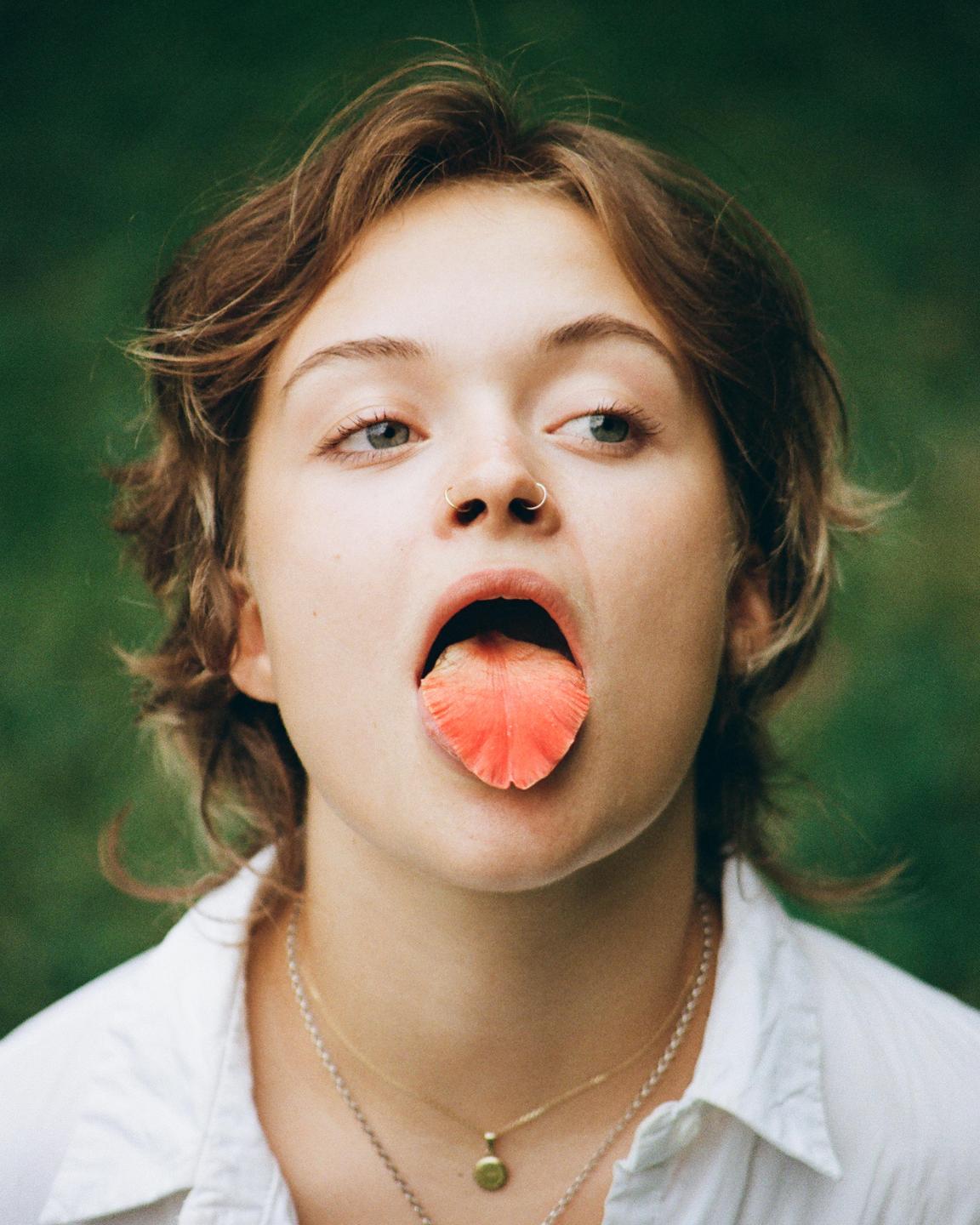 A woman with a flower petal on her tongue looks to the side.
