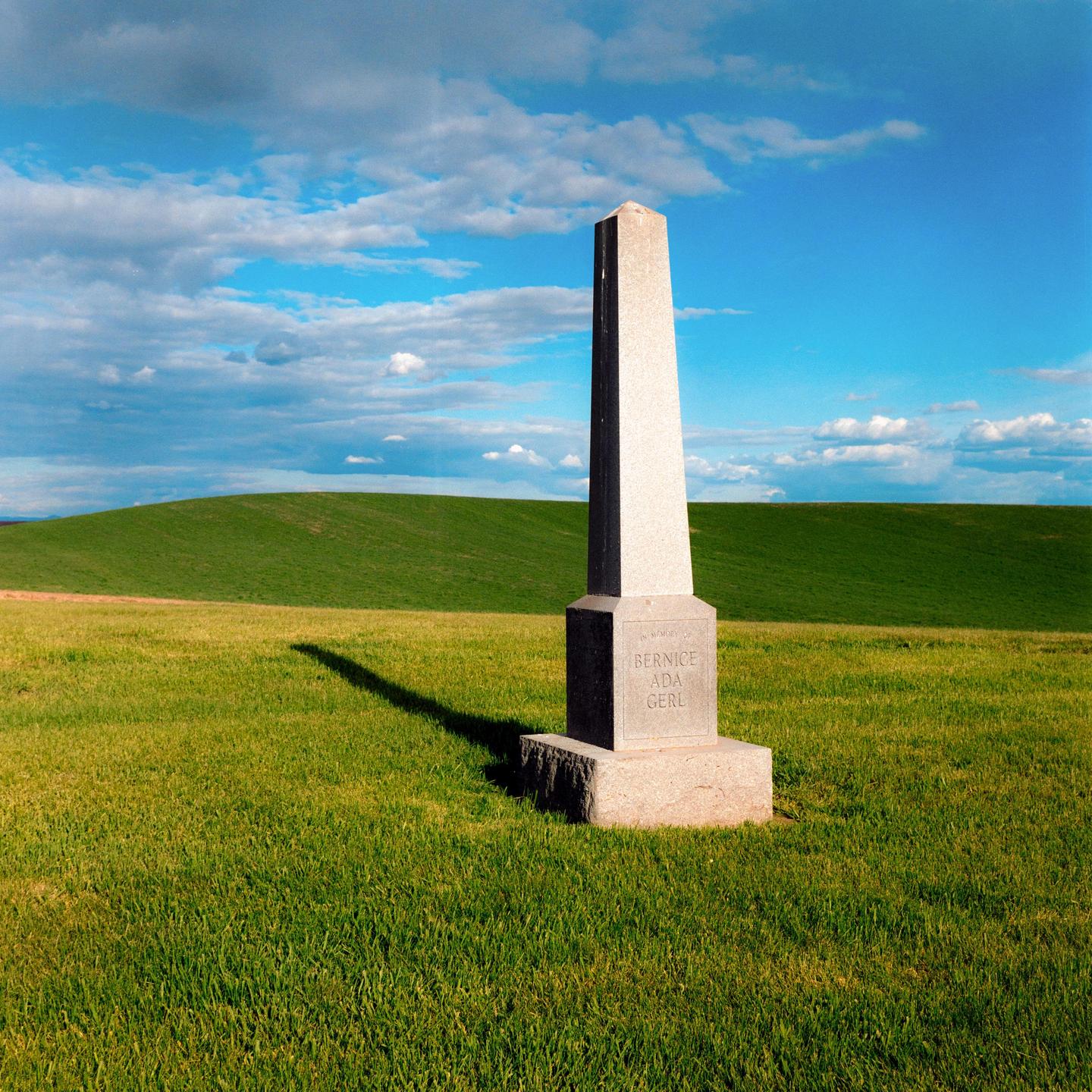 A tall, light-colored stone monument stands in a field of green grass under a blue sky with white clouds.