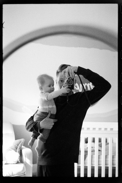 A man holds a baby while taking a mirror self-portrait in a room with a crib.
