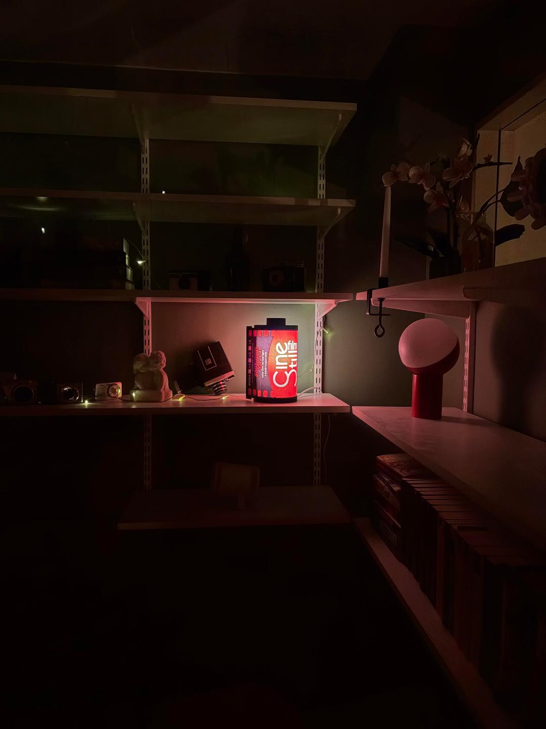 A red film canister lamp illuminates a shelf with various objects in a dark room.