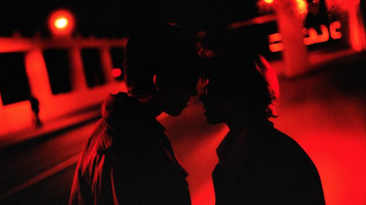 Two people are silhouetted against a red-lit background, facing each other closely.