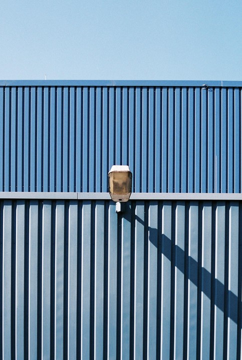 A street light is mounted on a blue corrugated metal wall under a clear sky.