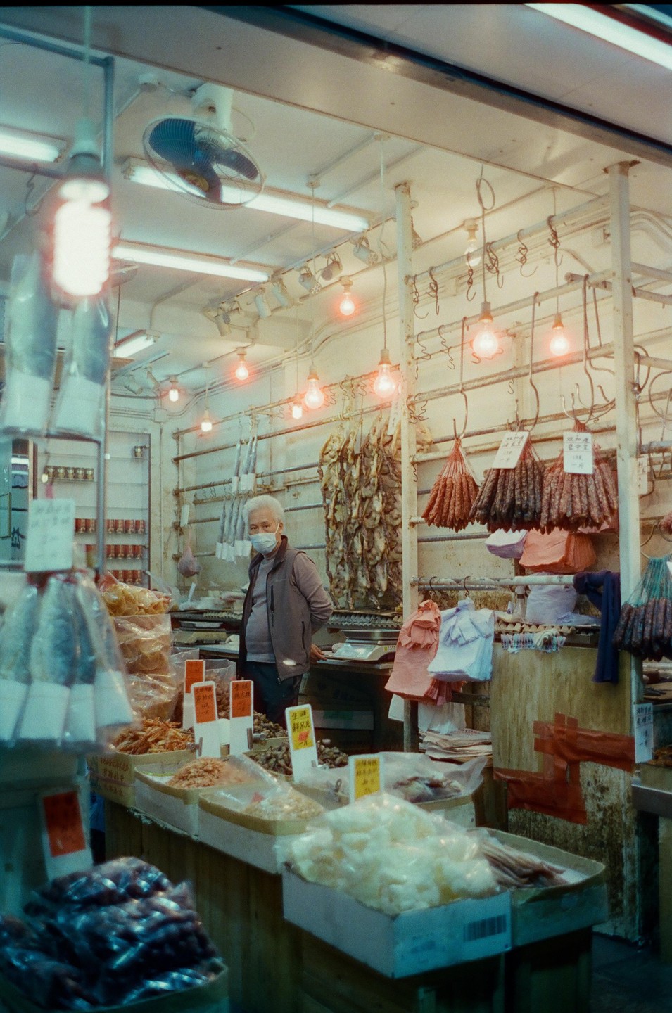 A man wearing a face mask stands behind a counter filled with various dried goods and sausages hanging from the ceiling in a market stall.
