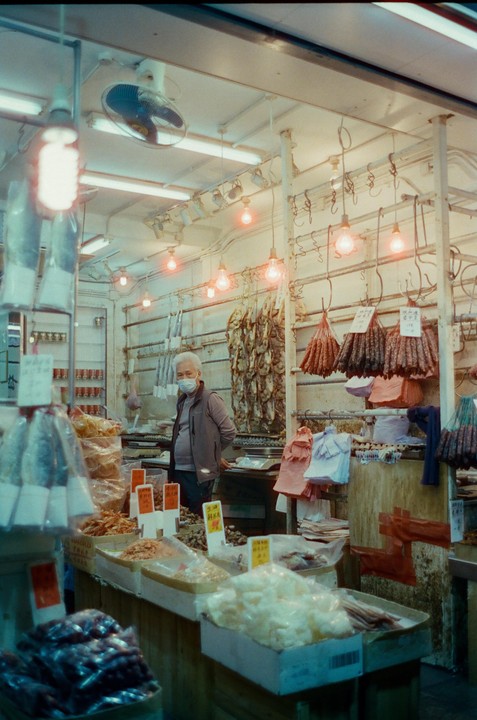 A man wearing a face mask stands behind a counter filled with various dried goods and sausages hanging from the ceiling in a market stall.