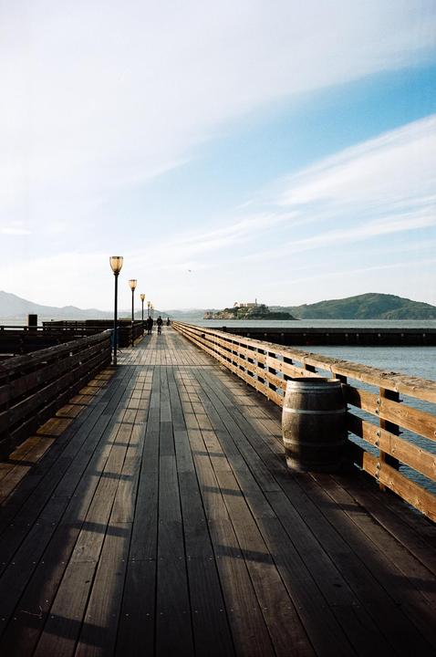 A long wooden pier with streetlights and a barrel extends into the water towards an island with a building on it.