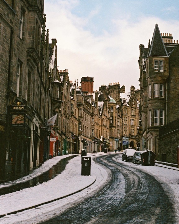 A snow-covered street curves through a historic city with tall stone buildings lining both sides.