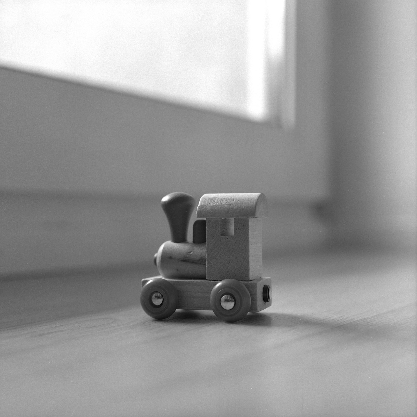 A wooden toy train sits on a wooden floor next to a window.